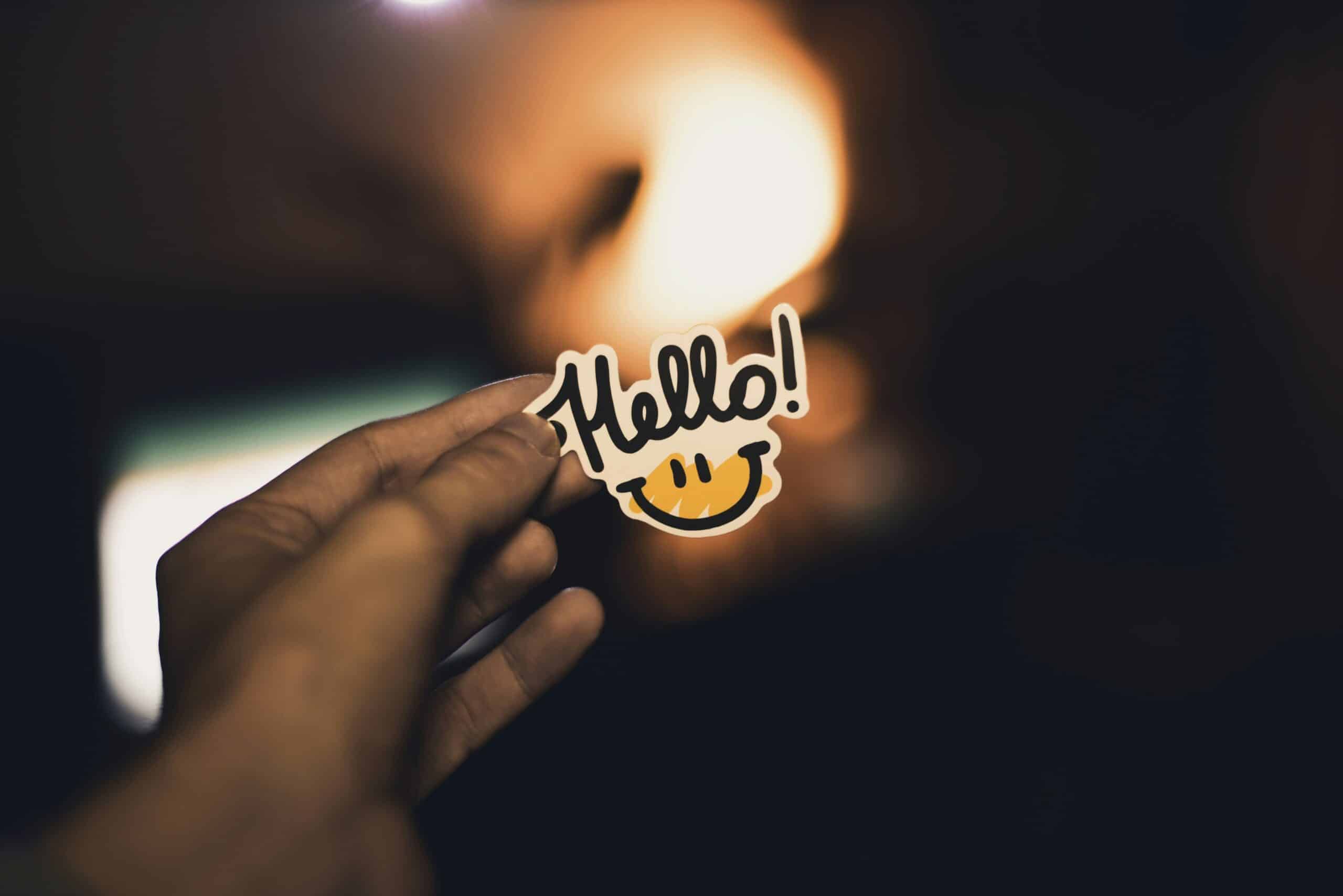 "Close-up of a hand holding a cheerful crafting sticker that says 'Hello!' in playful cursive, with a smiling yellow face below. Warm, blurred background adds a cozy, welcoming feel."