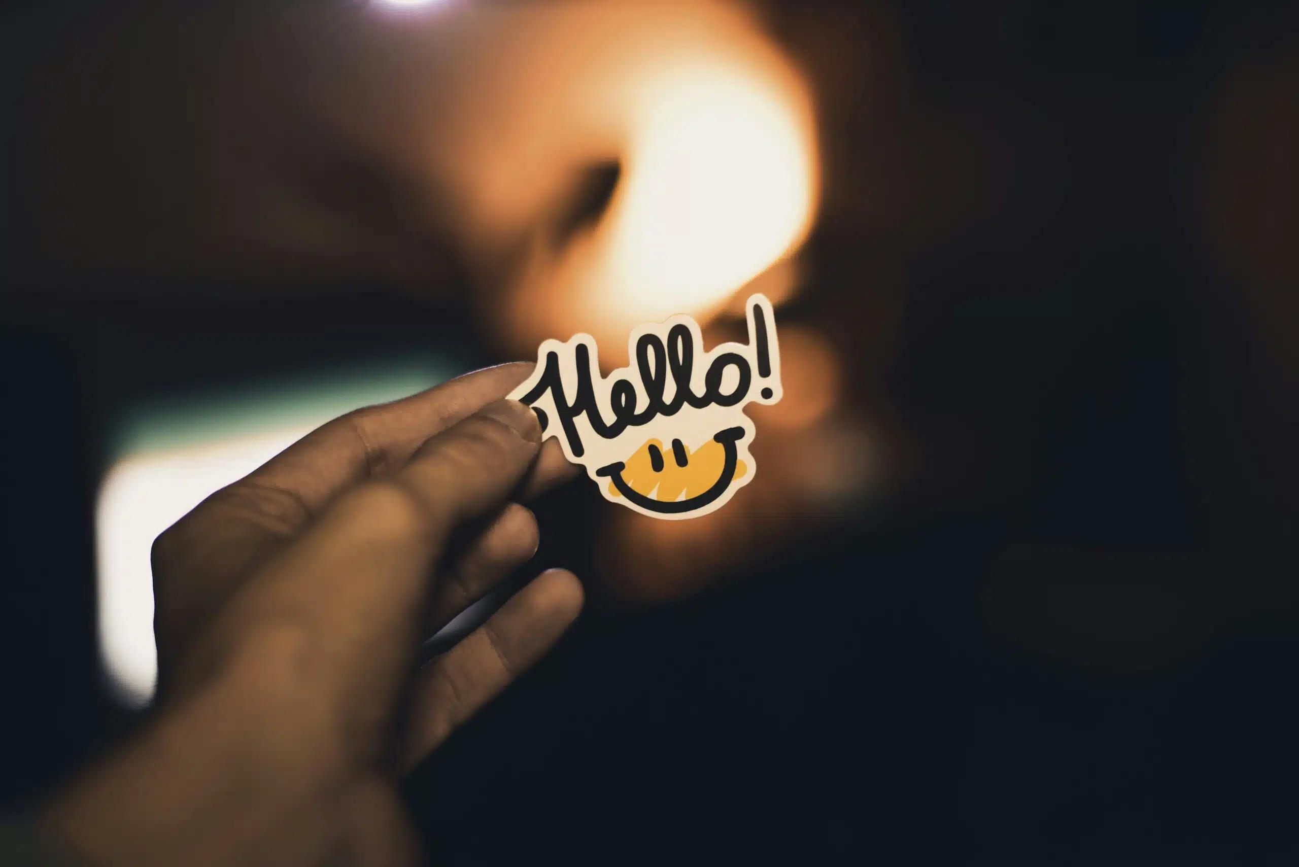 "Close-up of a hand holding a cheerful crafting sticker that says 'Hello!' in playful cursive, with a smiling yellow face below. Warm, blurred background adds a cozy, welcoming feel."